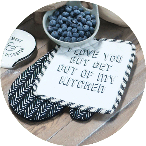 A black and white hot pad and mit set that says I Love You But Get Out of My Kitchen, with a bowl of blueberries and a spoon rest in the background.